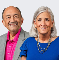 At left, a smiling older man in a grey jacket and a pink shirt. At right is an older woman with shoulder length grey hair in a turquoise top and wearing a beaded necklace.